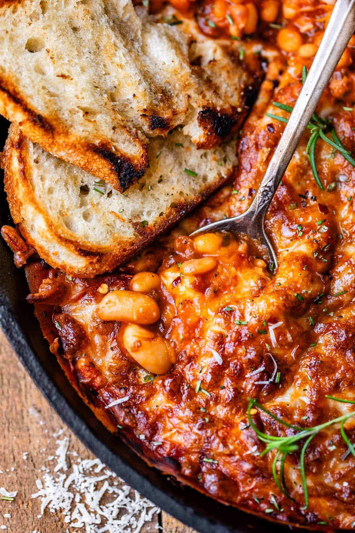 metal spoon and sourdough slice stuck into a cast iron skillet of cheesy beans.
