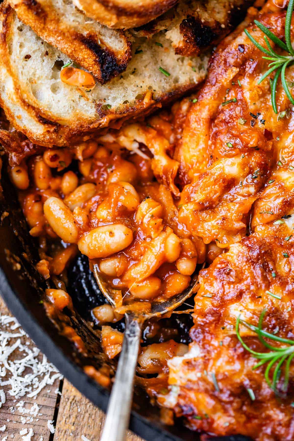 metal spoon and sourdough slice stuck into a cast iron skillet of cheesy beans.