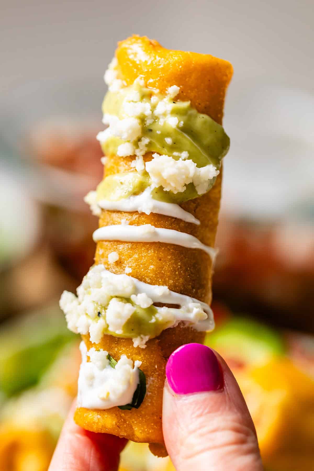 finger and thumb holding vertically a flauta topped with crema and cotija cheese.