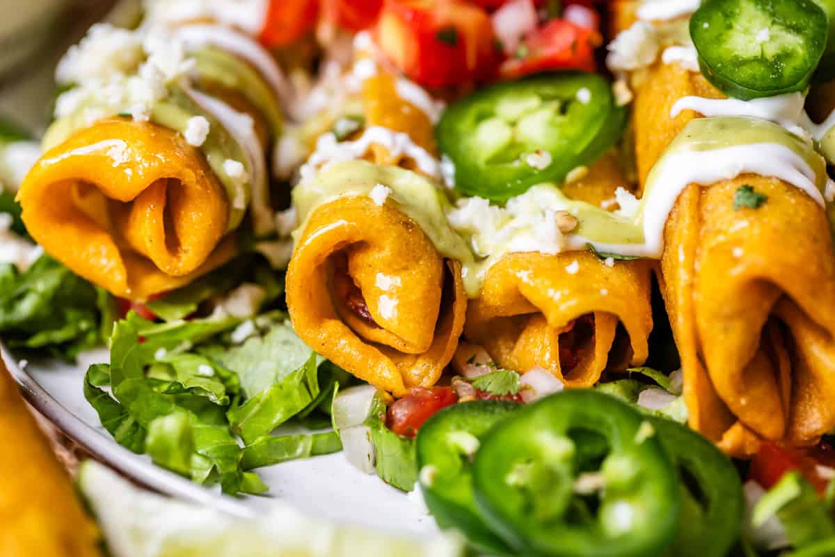 side view of 4 fried flautas on a bed of shredded lettuce with crema and other garnish.