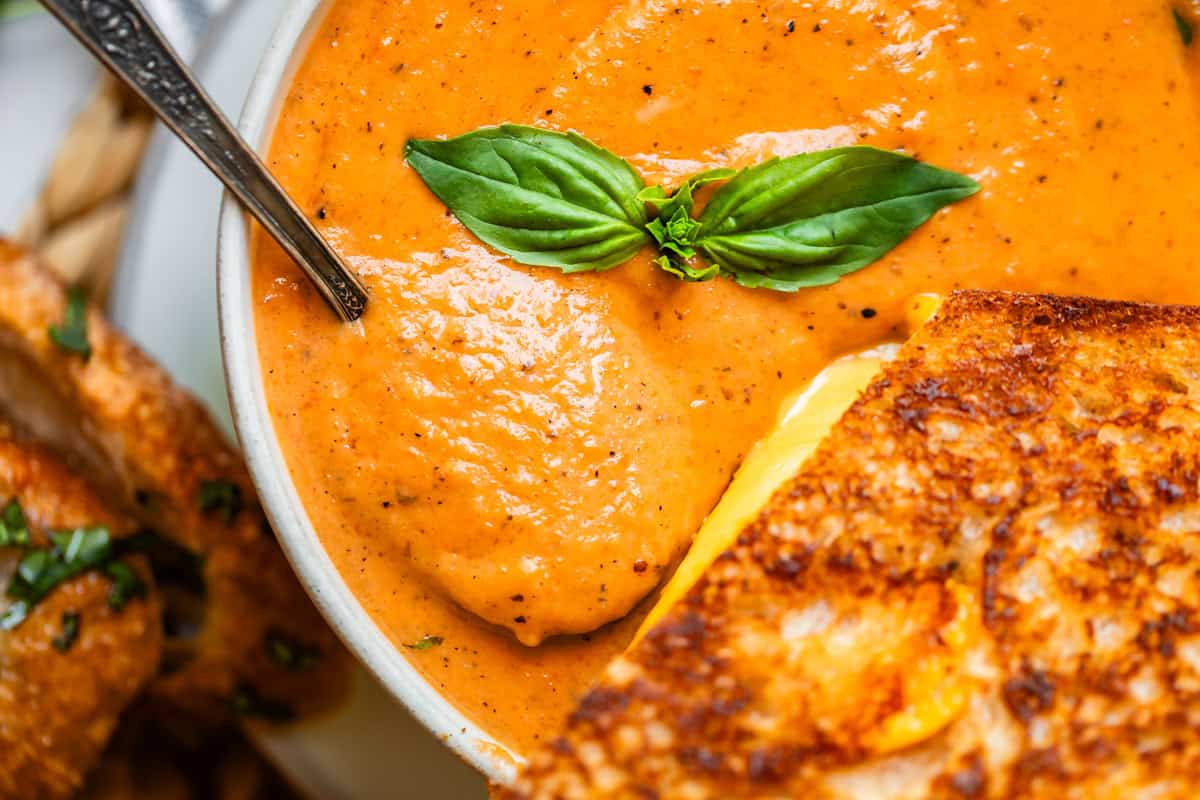 close up of bowl of tomato soup with metal spoon and grilled cheese in it.
