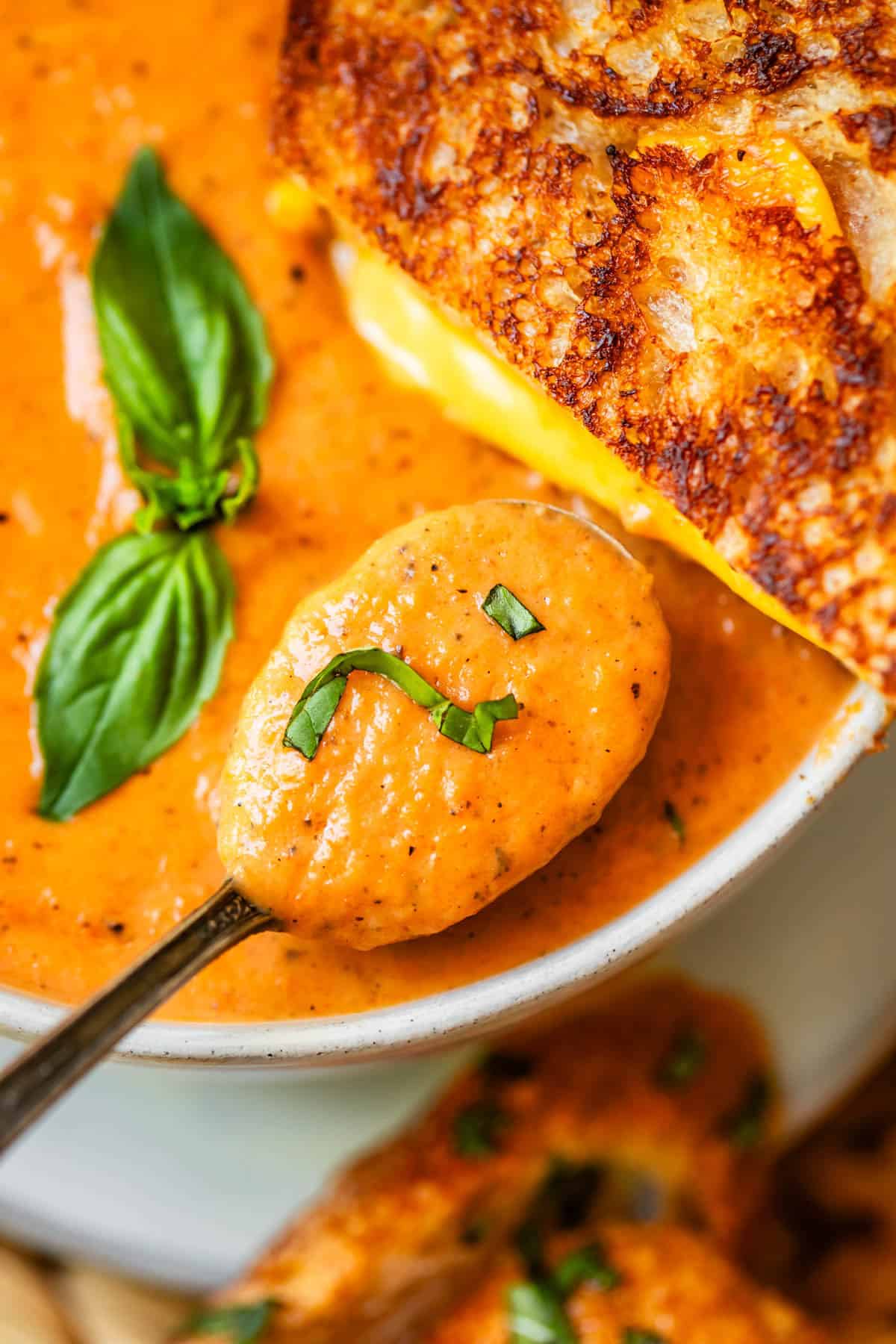 metal spoon holding a spoonful of tomato bisque with basil and a grilled cheese sandwich behind it.