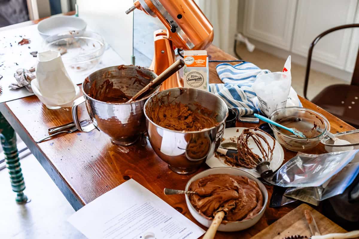 a messy kitchen counter with lots of chocolate frosting in various mixing bowls.