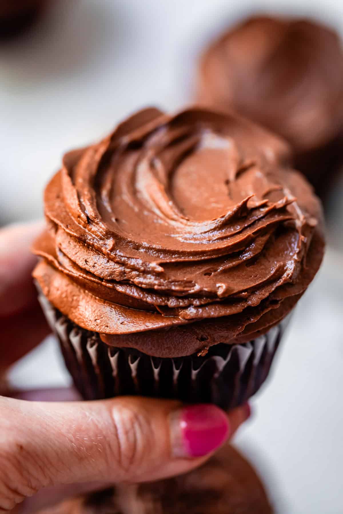 hand holding a chocolate cupcake in liner topped with chocolate frosting.