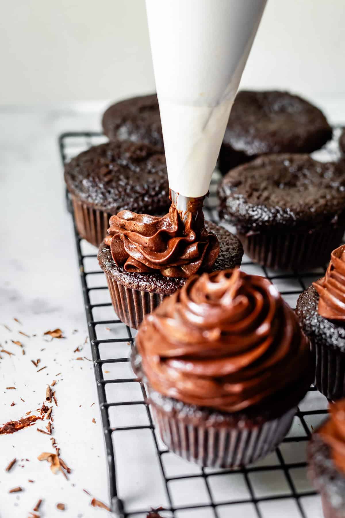 chocolate frosting being piped onto a chocolate cupcake sitting on a cooling rack.