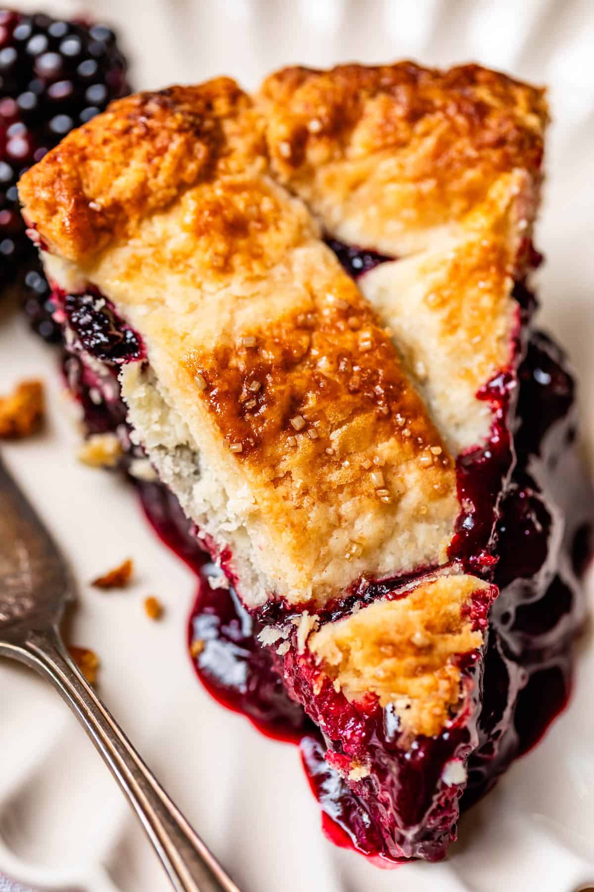 A close-up of a slice of blackberry pie with a golden, flaky crust and juicy, dark purple blackberry filling, served on a white plate with a fork beside it.