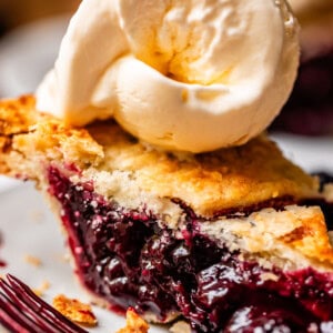 A close-up of a slice of blackberry pie with a golden, flaky crust, topped with a scoop of vanilla ice cream, on a white plate with a fork beside it.