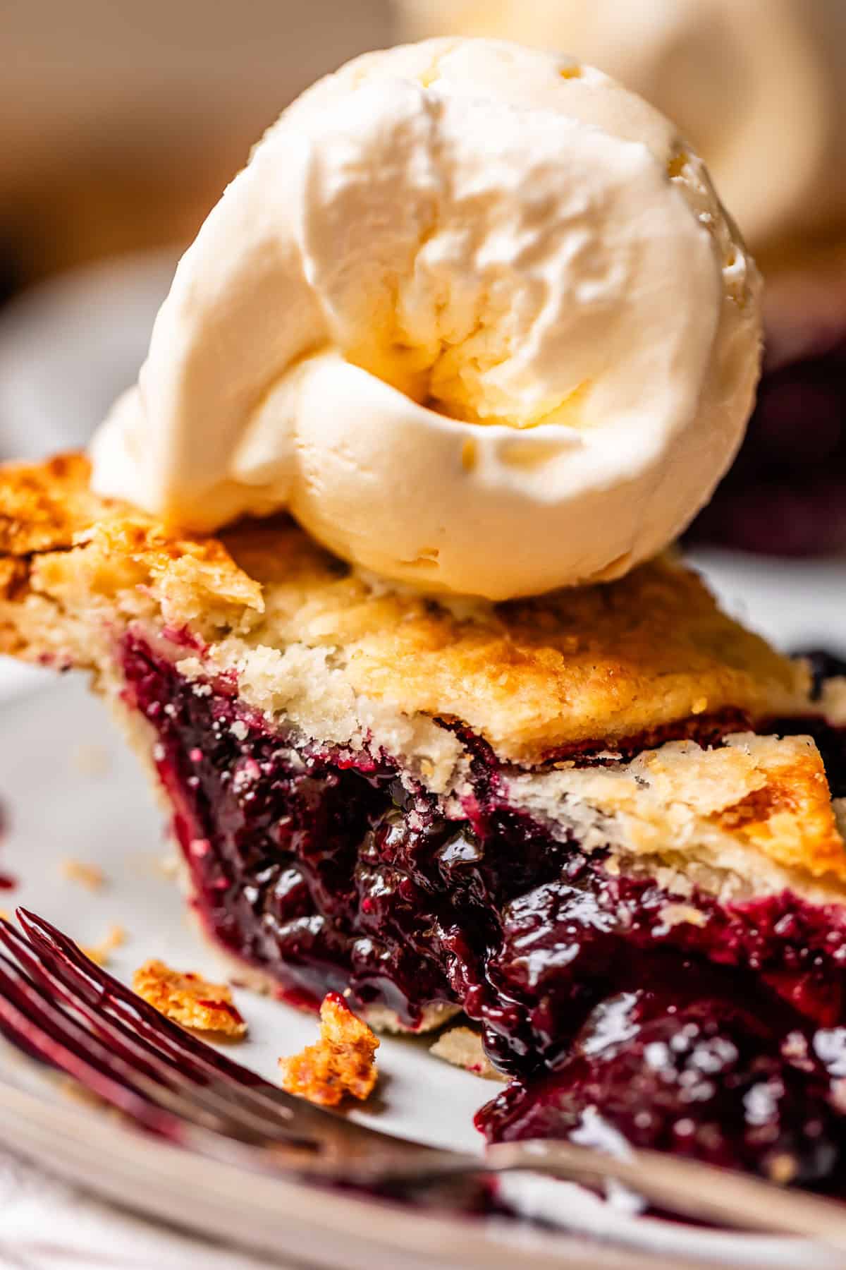 A close-up of a slice of blackberry pie with a golden, flaky crust, topped with a scoop of vanilla ice cream, on a white plate with a fork beside it.