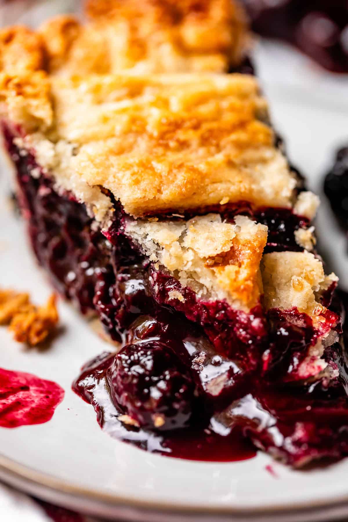 A close-up of a slice of berry pie with a golden, flaky crust and juicy, dark berry filling oozing onto a white plate.