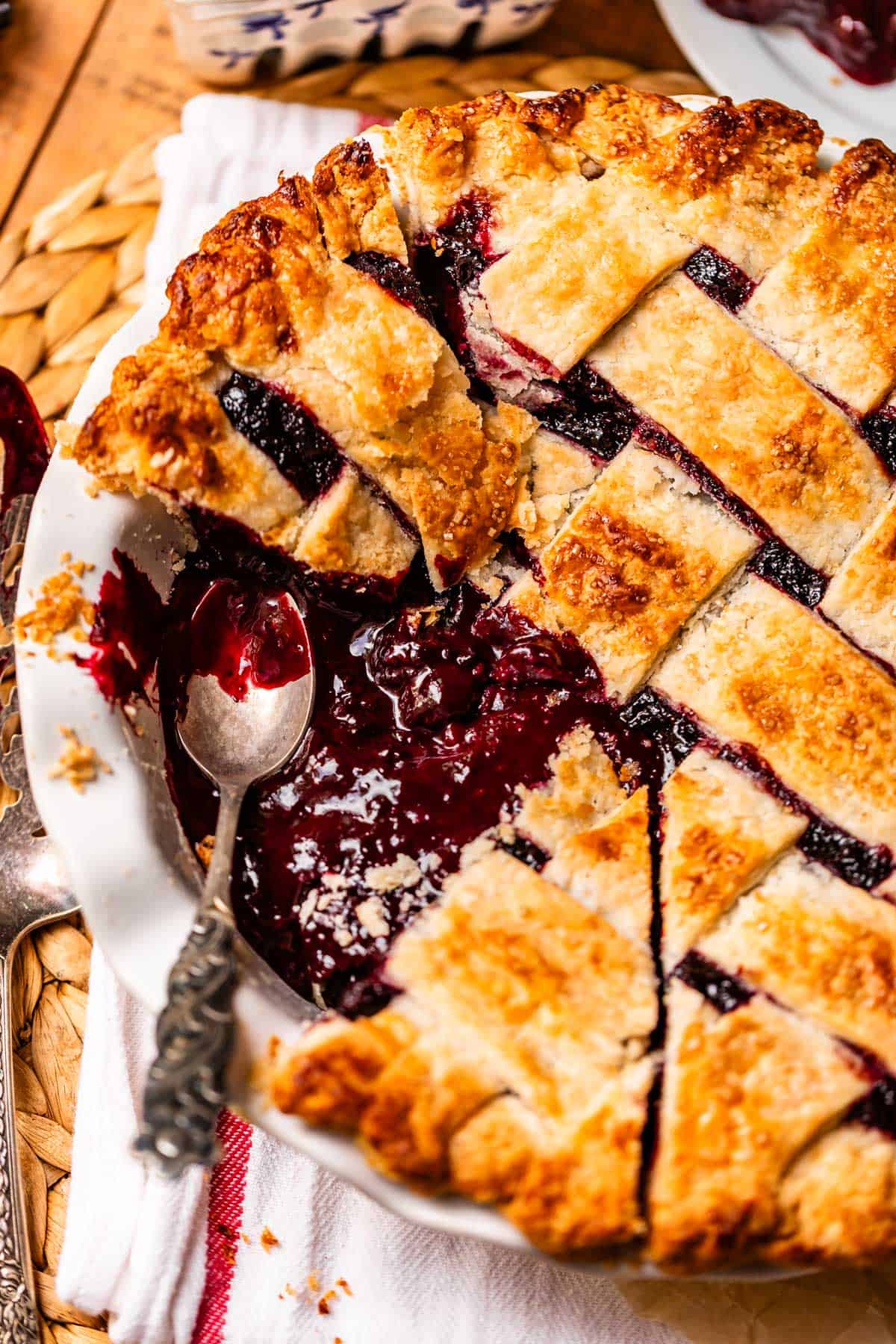 A close-up of a homemade blackberry pie with a golden lattice crust, partially sliced. A silver spoon rests in the pie dish, revealing the juicy berry filling. The pie sits on a white cloth with a woven placemat underneath.