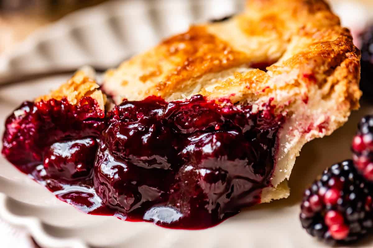 A close-up of a slice of blackberry pie on a white plate, showing thick, glossy blackberry filling and a golden, flaky crust, with a few fresh blackberries nearby.