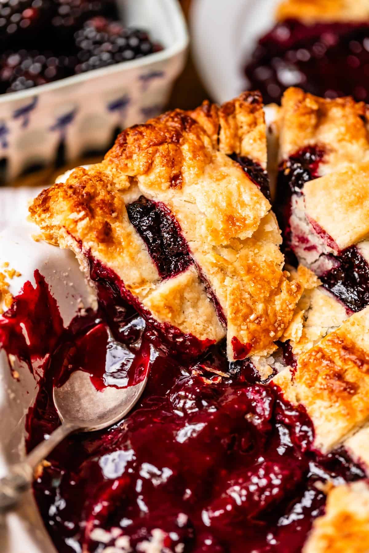 A close-up of a homemade blackberry pie with a golden, flaky lattice crust. A slice is being served, revealing juicy blackberry filling and a spoon resting in the pie dish. Fresh berries are seen in the background.