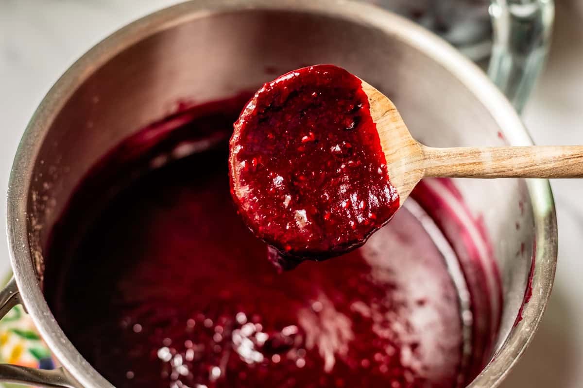 A wooden spoon holds up thick, dark red berry sauce over a saucepan filled with more of the same sauce.