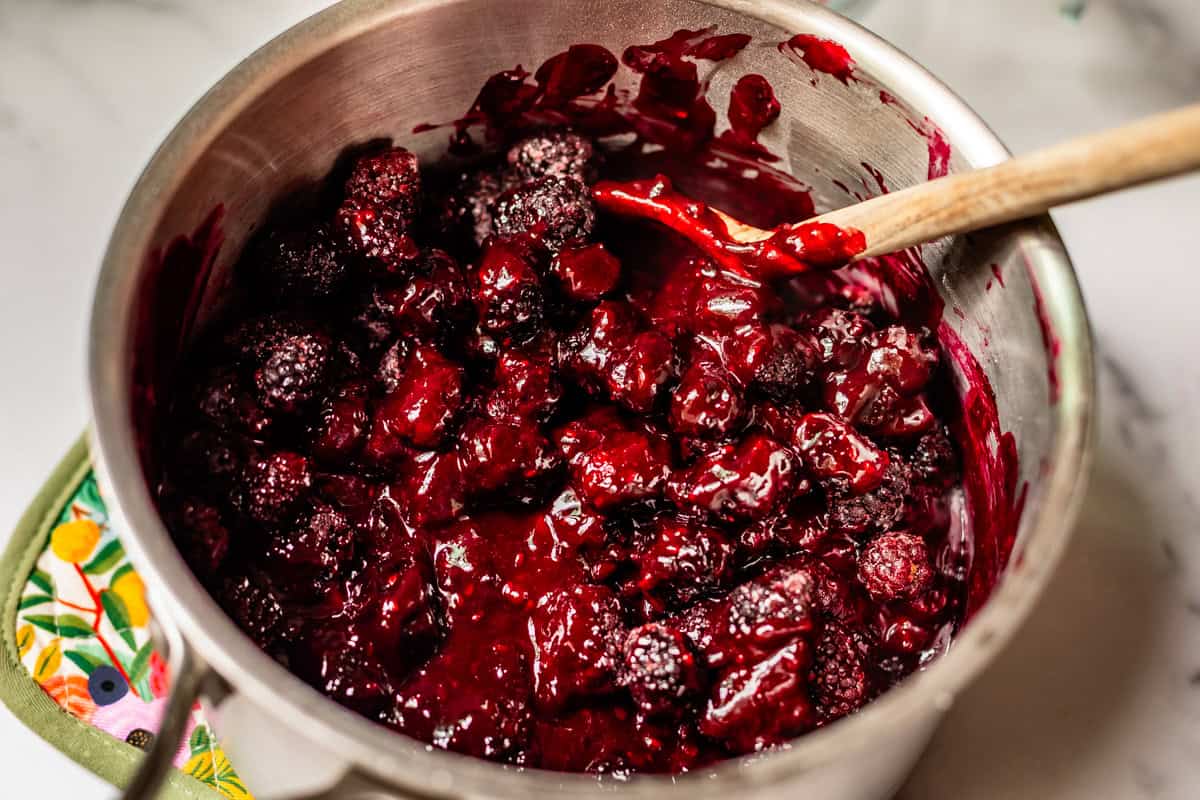 A stainless steel saucepan filled with thick, glossy mixed berry compote, with whole blackberries visible, being stirred by a wooden spoon on a white countertop.