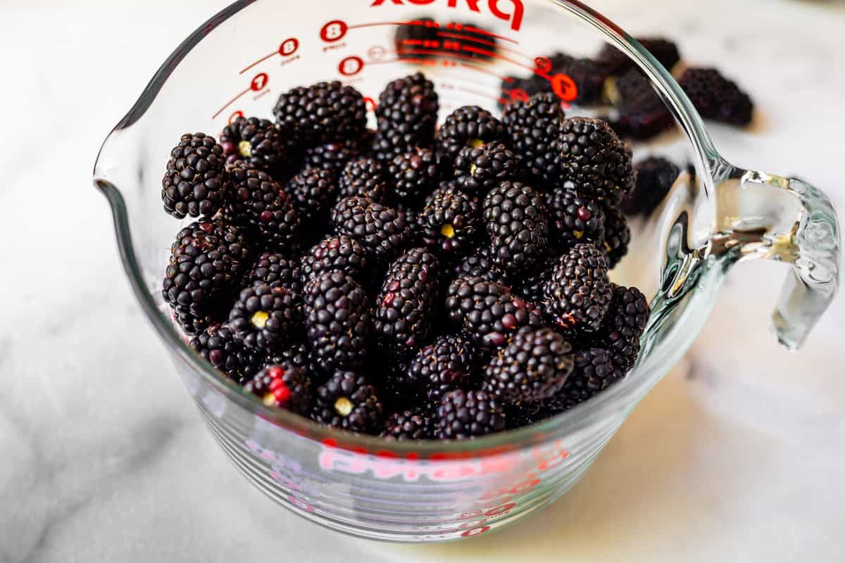 A glass measuring cup filled with fresh blackberries sits on a white surface, with a few blackberries scattered in the background.