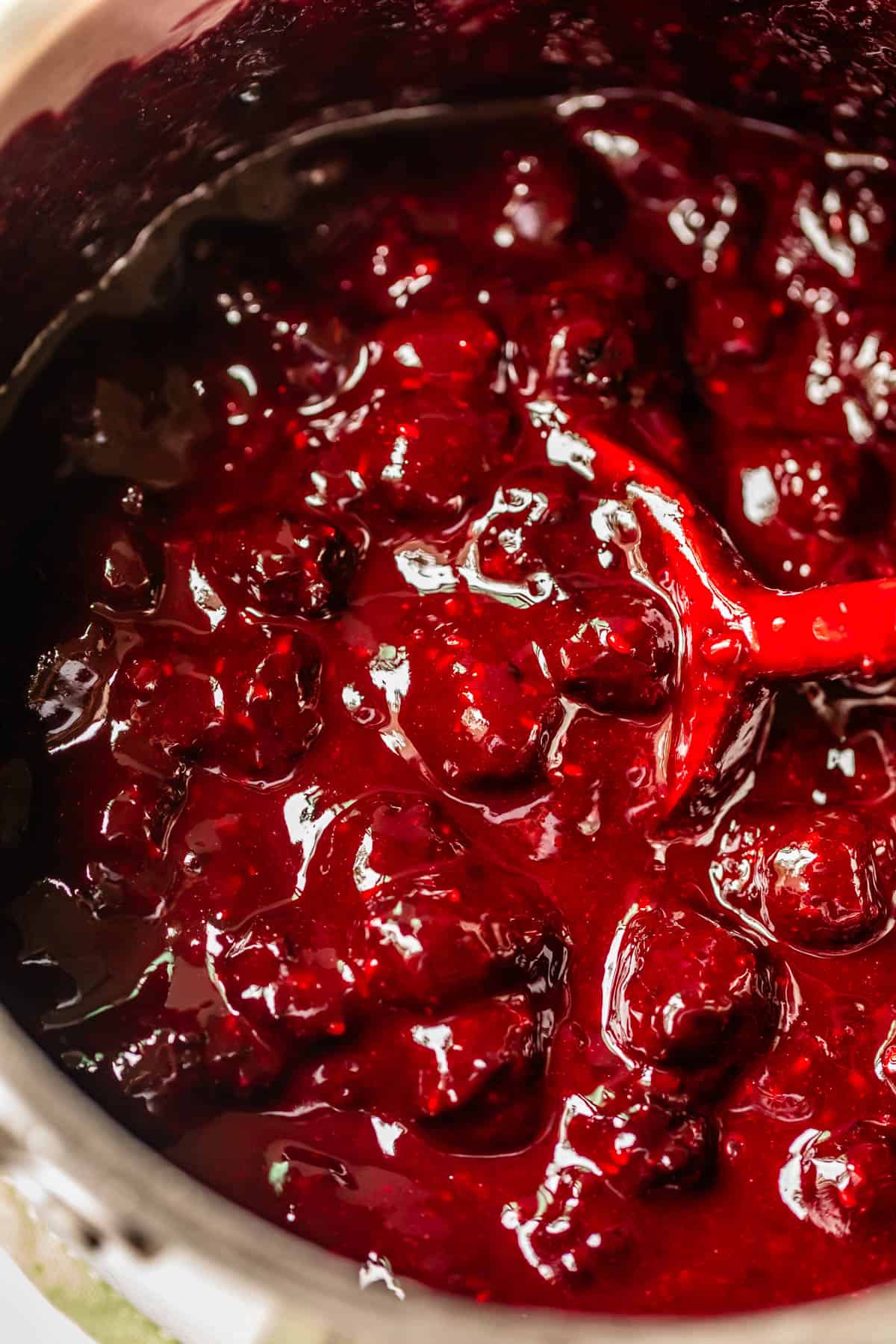Close-up of a pot filled with thick, glossy blackberry pie filling, with whole berries and juices visible. A red spoon is partially submerged, stirring the mixture.