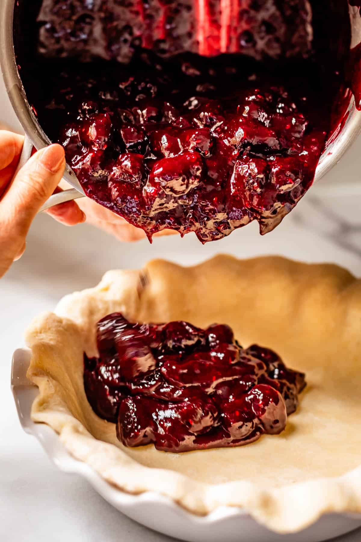 A person pours thick blackberry pie filling from a saucepan into an unbaked pie crust in a ceramic pie dish.
