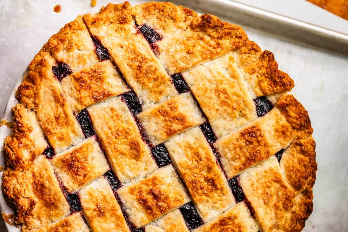 A freshly baked pie with a golden-brown, sugar-coated lattice crust, revealing dark berry filling underneath, sits on a metal baking sheet.