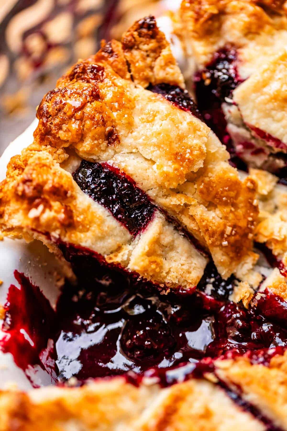 A close-up of a slice of blackberry pie with a golden, flaky lattice crust and juicy, dark berry filling spilling out onto the dish.