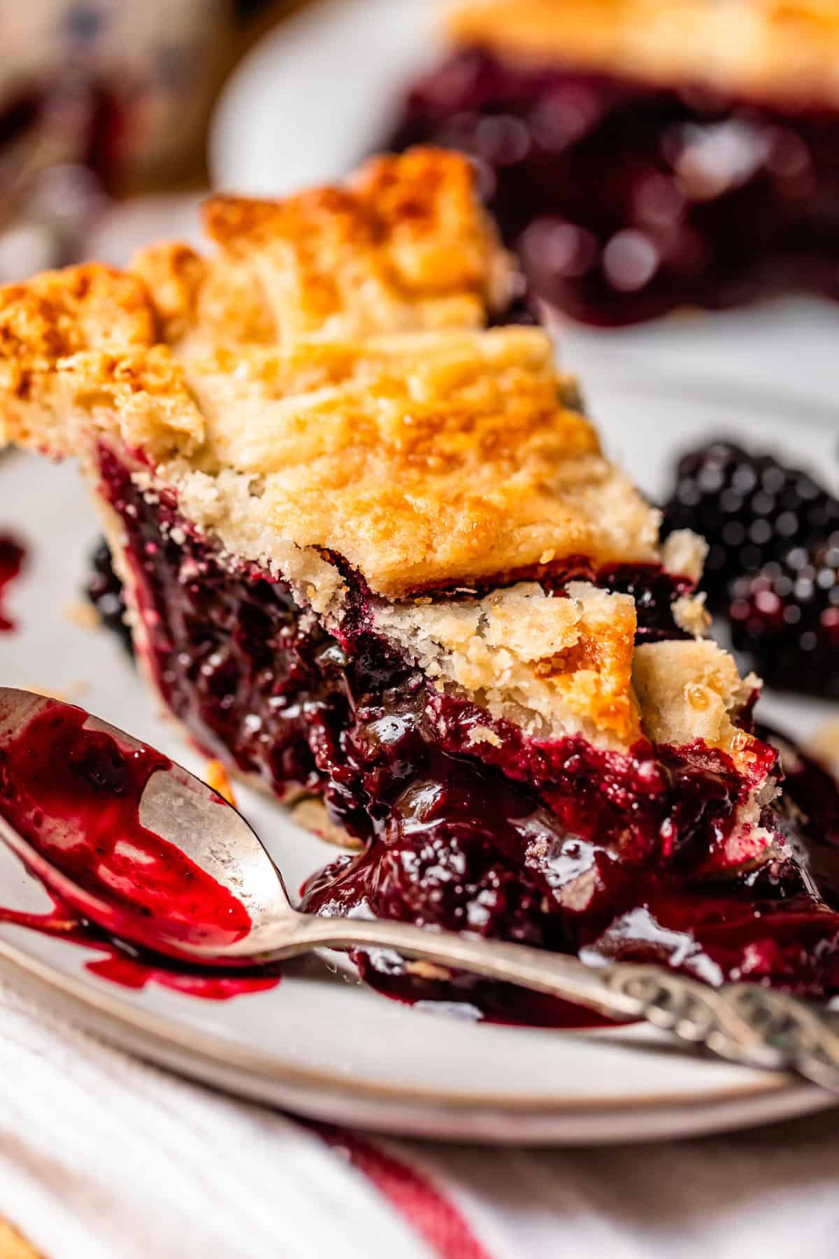 A close-up of a slice of blackberry pie on a white plate, with a golden, flaky crust and juicy, dark purple blackberry filling oozing out. A spoon with pie filling rests next to the slice.