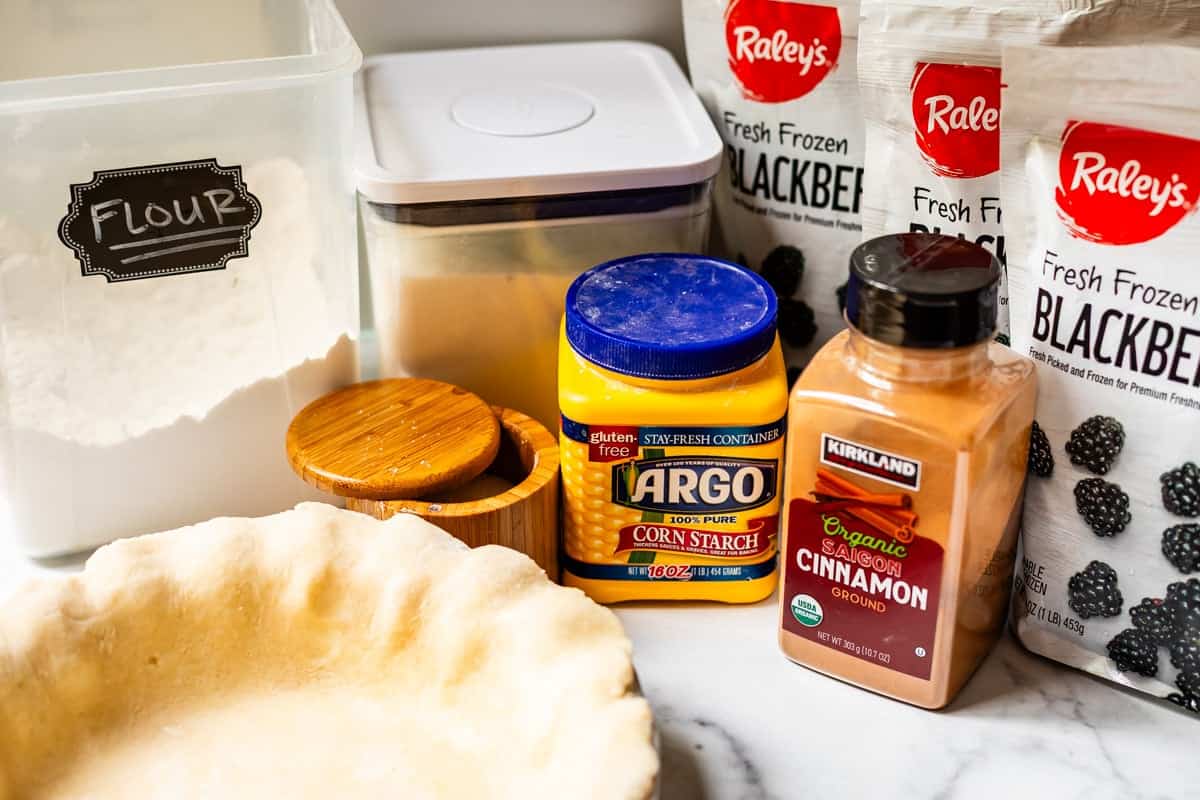 A marble counter holds a pie crust, a flour container, a sugar canister, a wooden container, cornstarch, cinnamon, and bags of frozen blackberries, all set for baking.