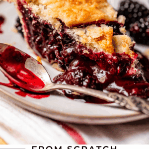 A close-up of a slice of blackberry pie with a golden, flaky crust and juicy blackberry filling on a plate, with a fork and fresh blackberries beside it. Text reads From Scratch Blackberry Pie – The Food Charlatan.
