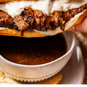 A hand holds a French dip sandwich with melted cheese and shredded beef, above a bowl of au jus dipping sauce. Text below reads French Dip Sandwiches - The Food Charlatan.