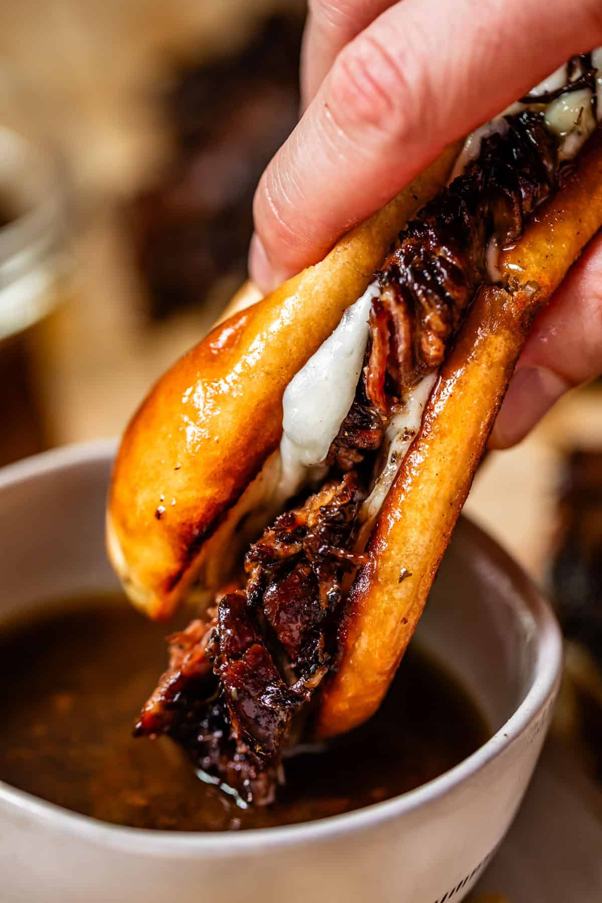 A hand dips a juicy, grilled beef sandwich with melted cheese into a bowl of brown au jus sauce. The sandwich is packed with tender meat and served on a golden, toasted bun.