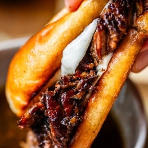 A close-up of a hand holding a French dip sandwich filled with juicy, shredded beef and melted cheese, with a bowl of au jus dipping sauce underneath.