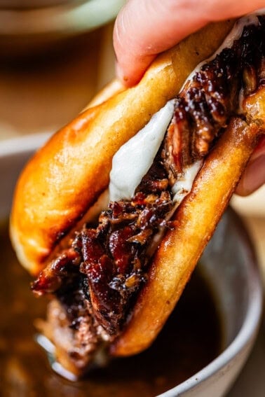 A close-up of a hand holding a French dip sandwich filled with juicy, shredded beef and melted cheese, with a bowl of au jus dipping sauce underneath.