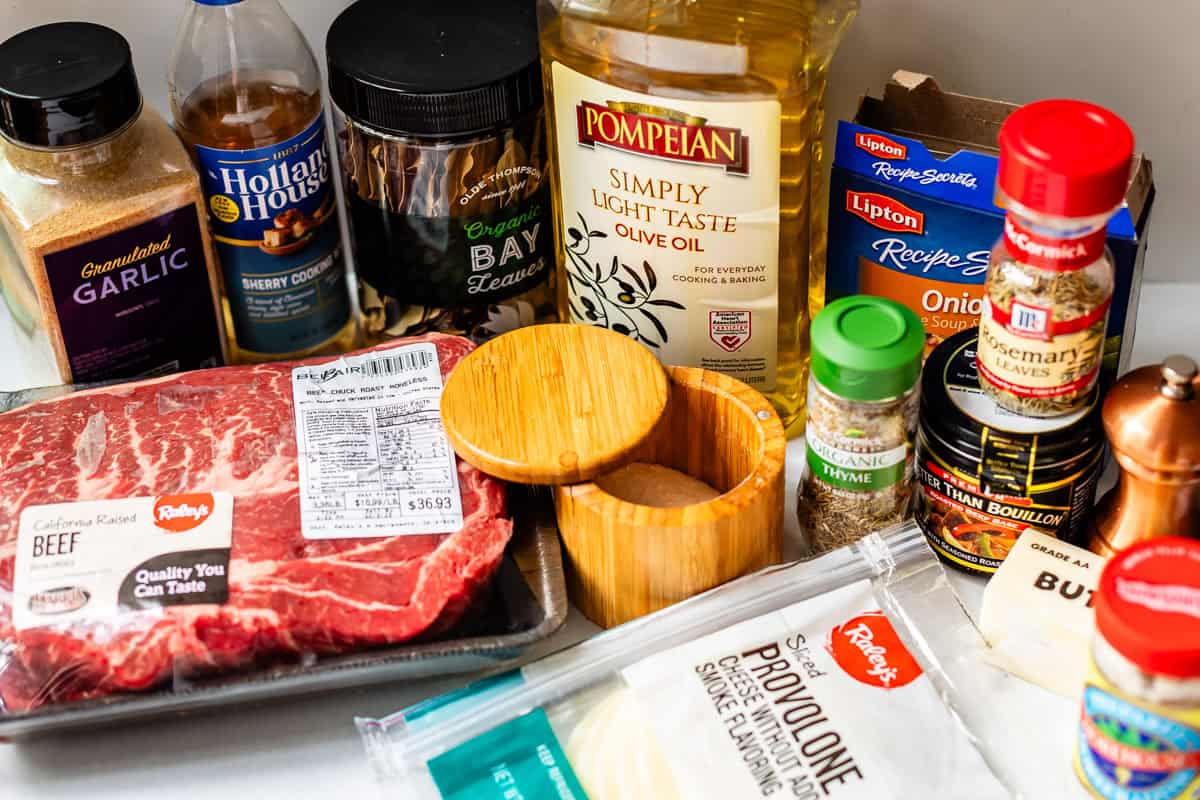 A variety of ingredients on a counter, including a beef roast, provolone cheese slices, olive oil, seasonings, beef bouillon, bay leaves, onion soup mix, sherry cooking wine, and spice containers.