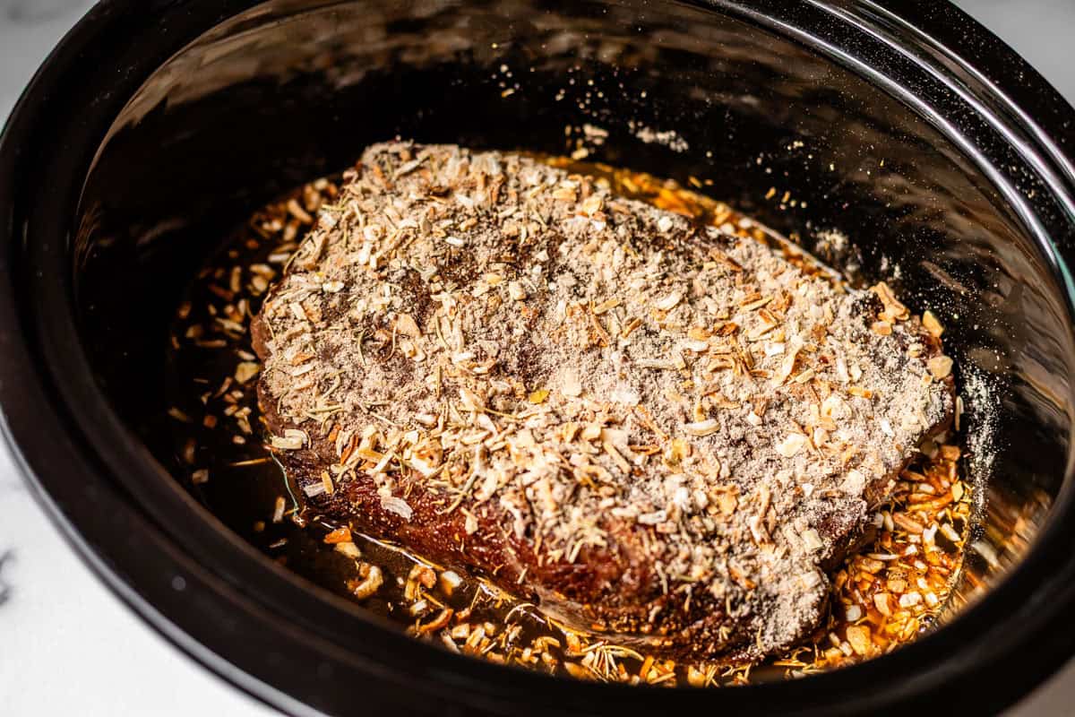 A seasoned beef roast sits in a slow cooker with visible dried onion soup mix and broth surrounding the meat, preparing to be cooked.