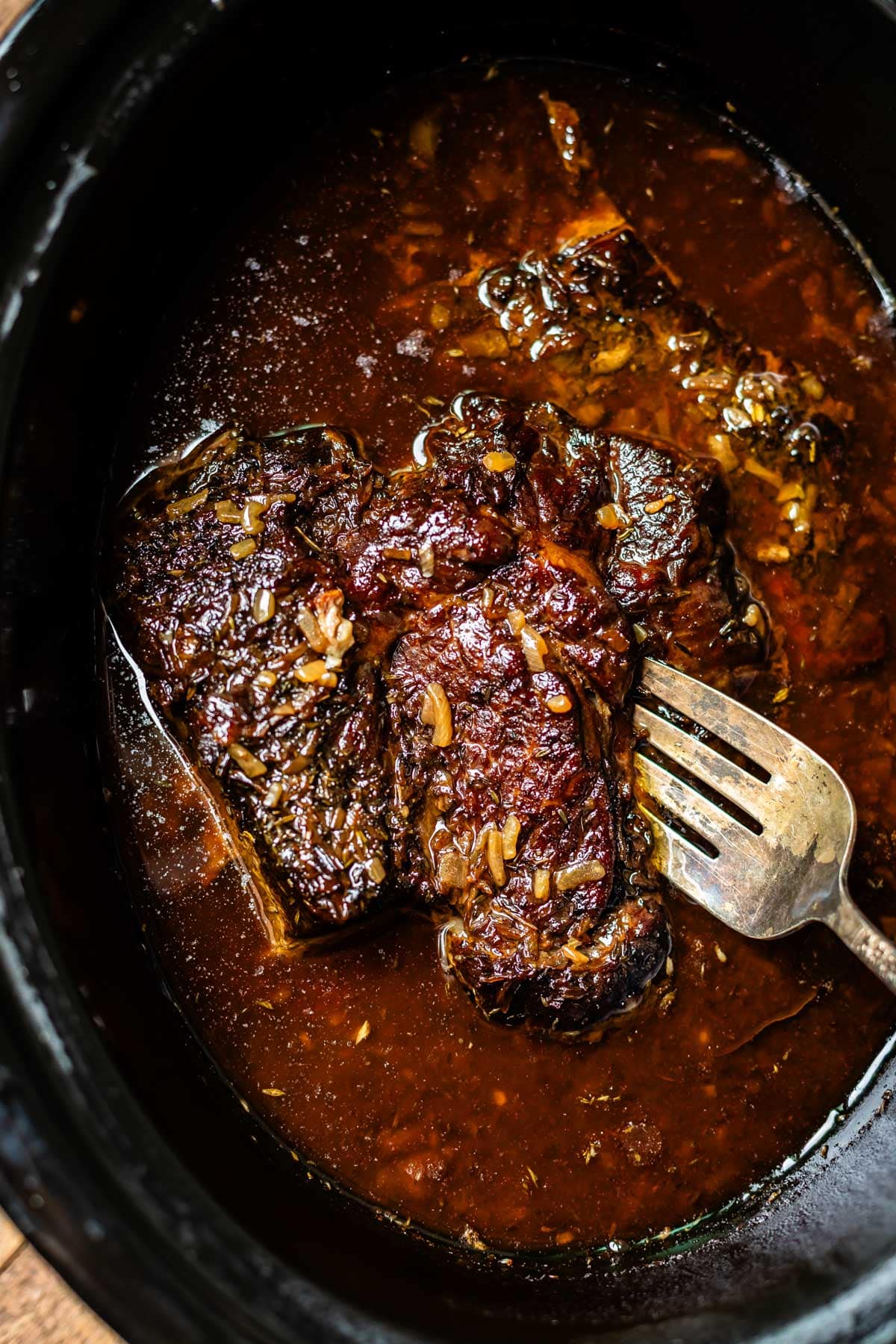 A close-up of a cooked beef roast in a slow cooker, sitting in a rich, dark brown sauce with visible seasonings. A metal fork rests beside the tender, juicy meat.