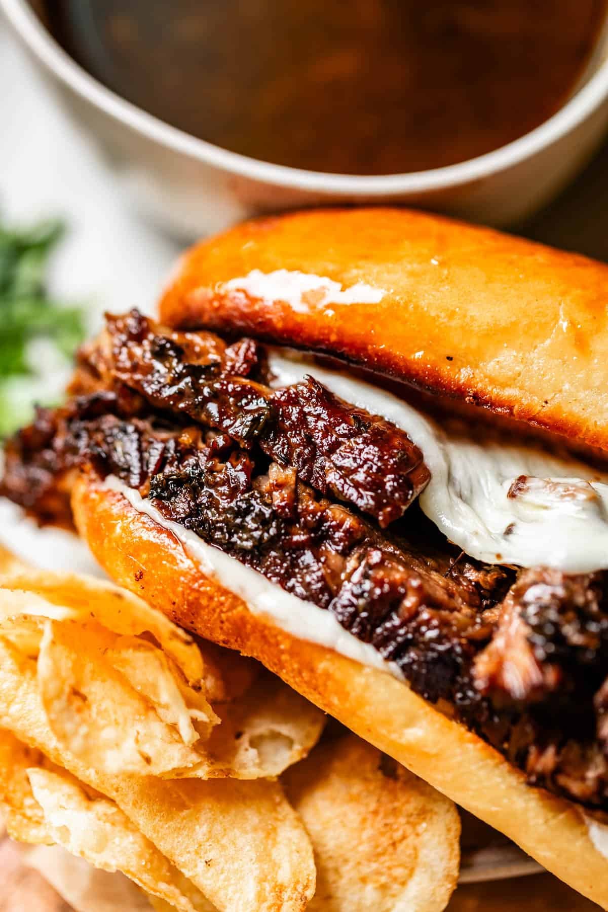 A close-up of a French dip sandwich with tender beef and melted cheese in a toasted bun, served with potato chips and a cup of au jus dipping sauce in the background.