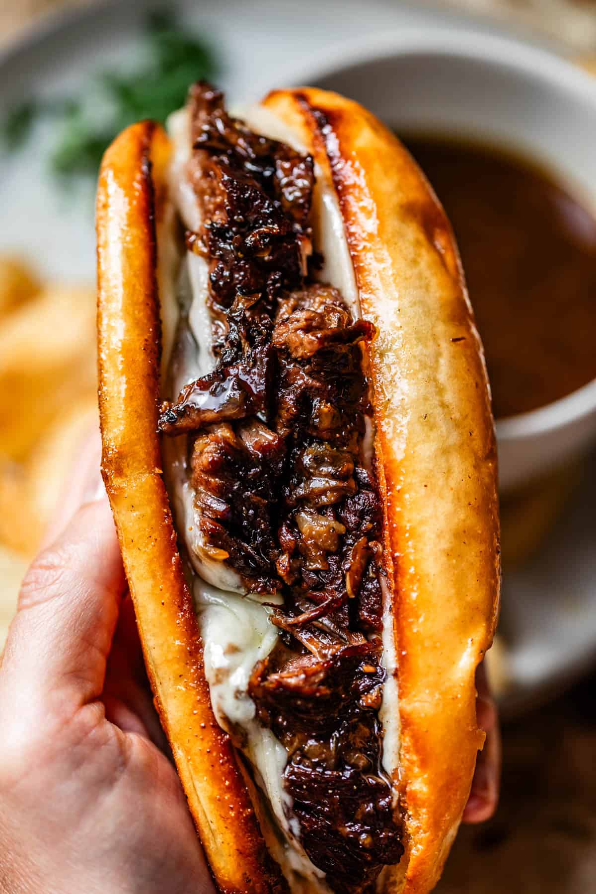 A close-up of a hand holding a toasted sandwich filled with melted cheese and tender, juicy pieces of beef, with a cup of au jus sauce blurred in the background.