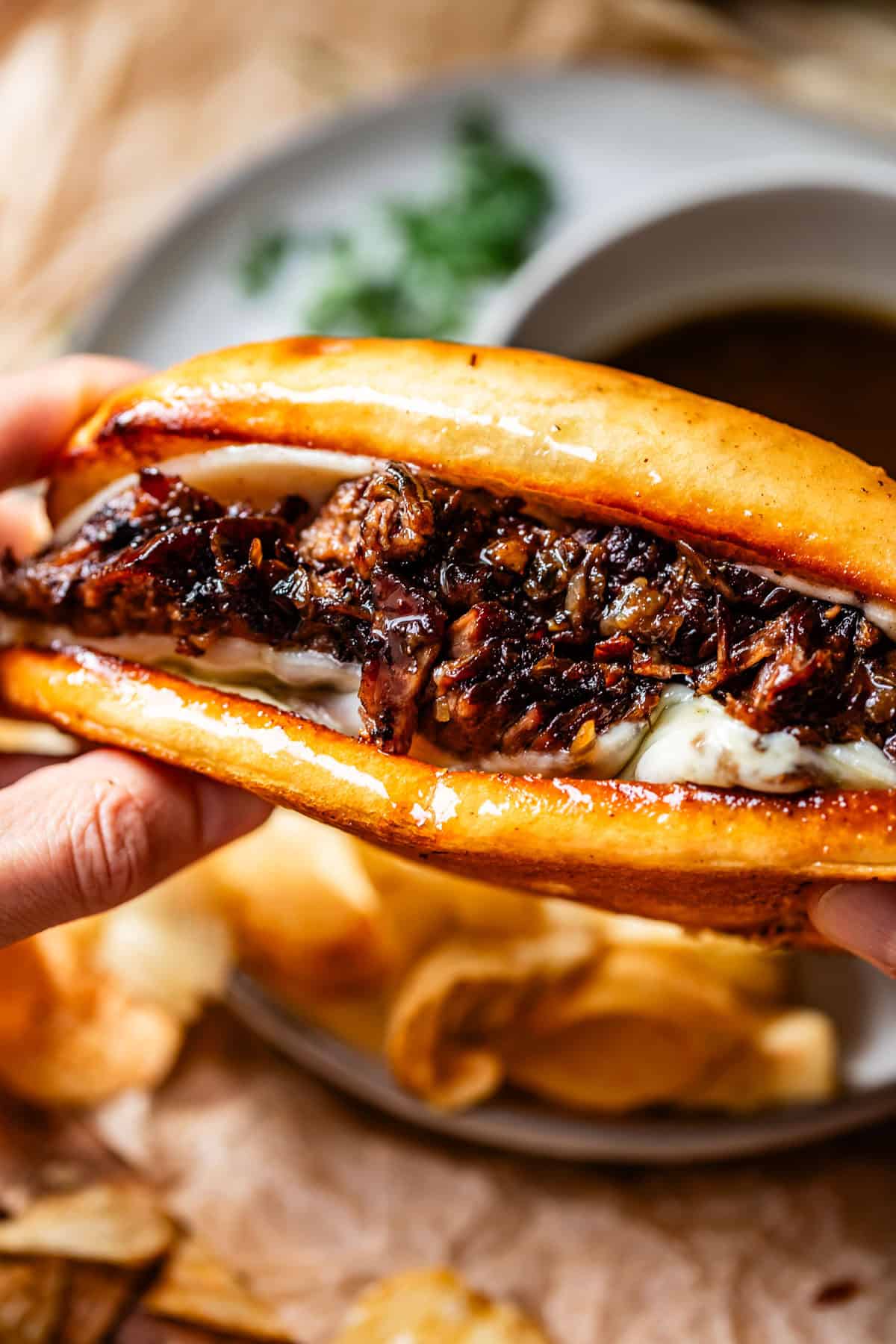 A close-up of hands holding a French dip sandwich filled with sliced beef and melted cheese, with a side of potato chips and a bowl of au jus in the background.