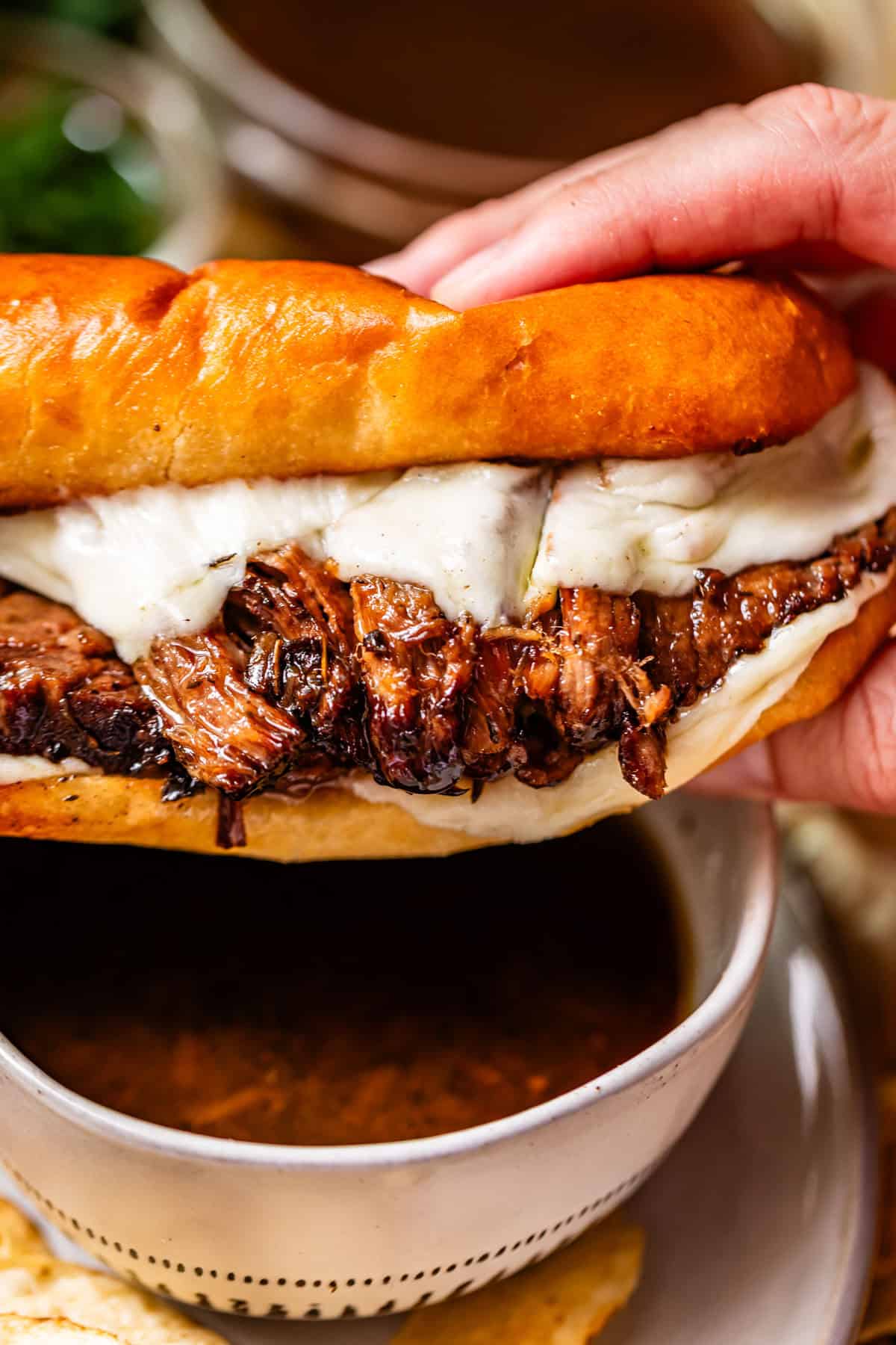 A hand holds a sandwich filled with shredded roast beef and melted cheese on a toasted bun, with a small bowl of au jus dipping sauce underneath.