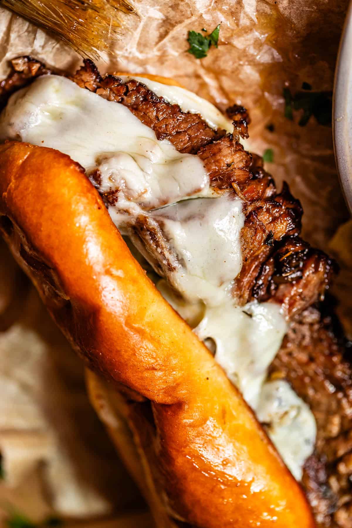 Close-up of a toasted sandwich roll filled with grilled steak and melted cheese, resting on brown parchment paper with parsley sprinkled around.