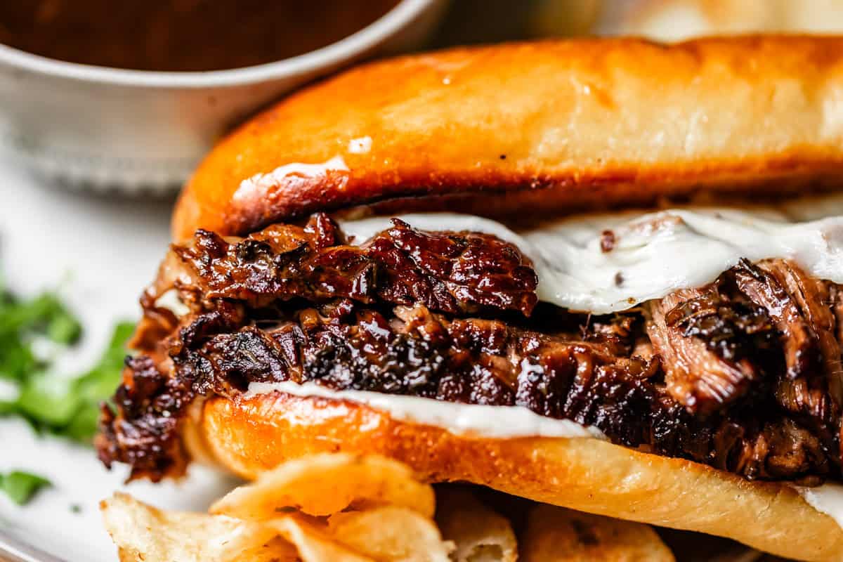 Close-up of a sandwich filled with shredded, juicy beef and melted cheese on a toasted bun, served with potato chips and a cup of dipping sauce in the background.
