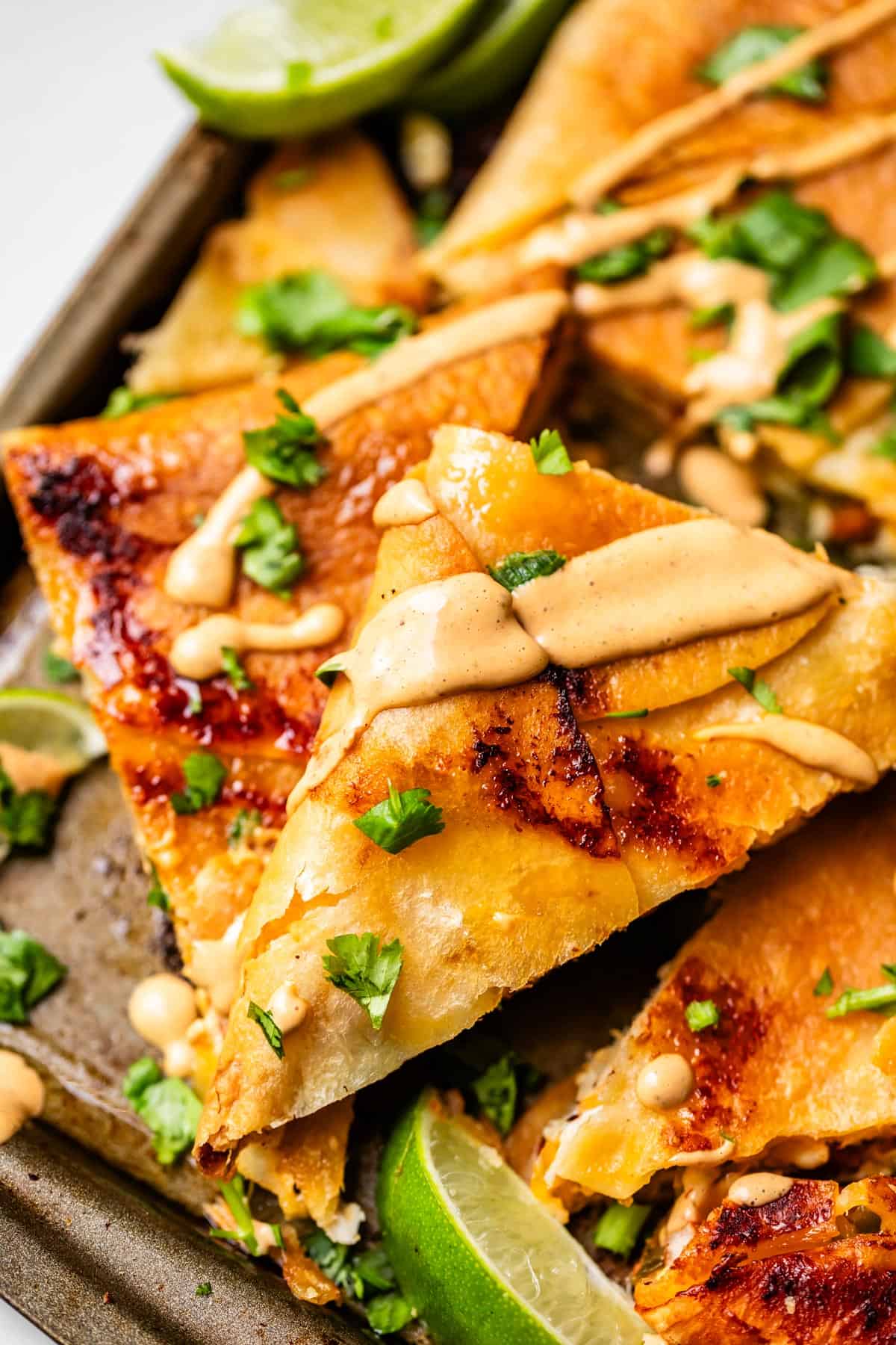 Close-up of crispy quesadilla triangles topped with chopped cilantro and a drizzle of creamy sauce, with lime wedges on the side.