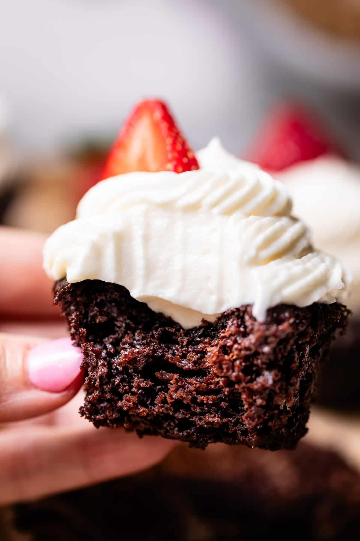 hand holding a chocolate cupcake with white frosting and a strawberry slice.