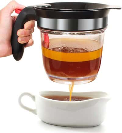 A hand holds a grease separator with strained liquid pouring into a white gravy boat.