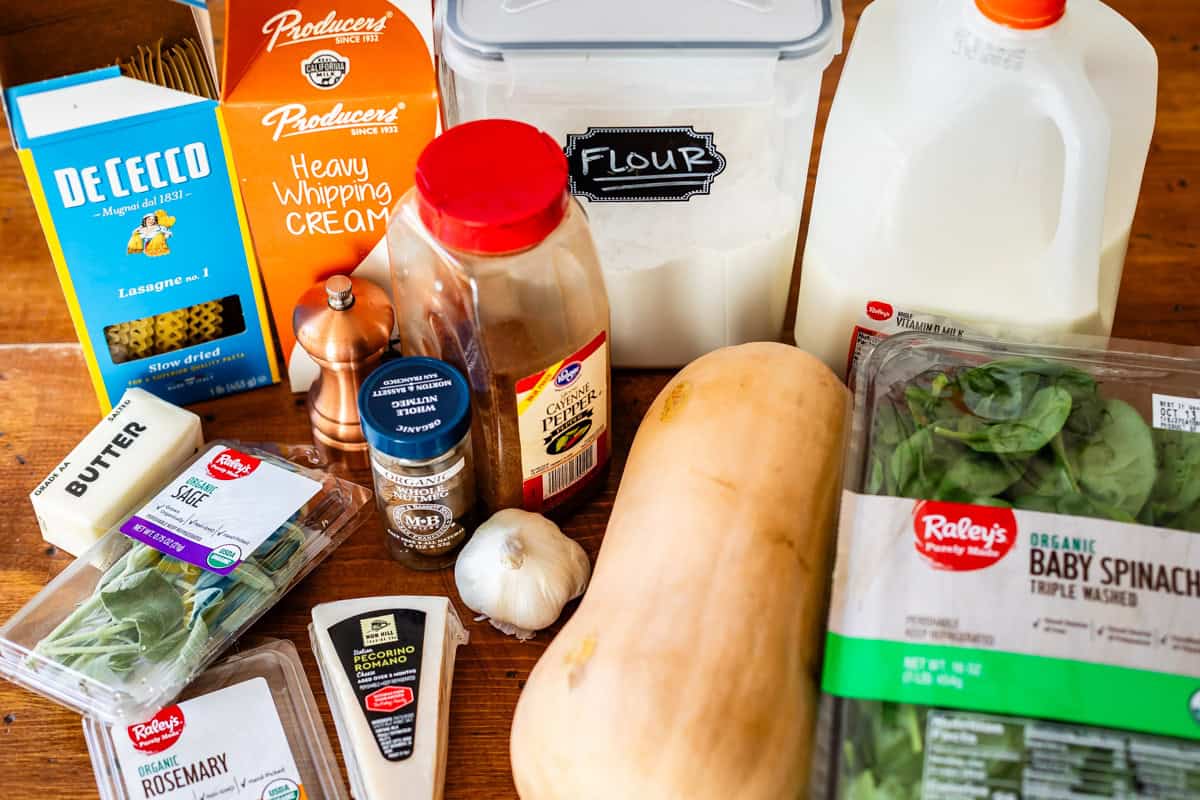 A variety of ingredients on a wooden surface, including lasagna noodles, heavy cream, flour, milk, butter, garlic, baby spinach, rosemary, pepper, nutmeg, a block of cheese, and a butternut squash.