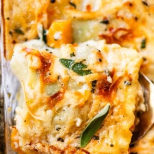 A close-up of a cheesy layered casserole with golden-brown edges, melted cheese, visible herbs, and a serving being lifted out with a spatula.