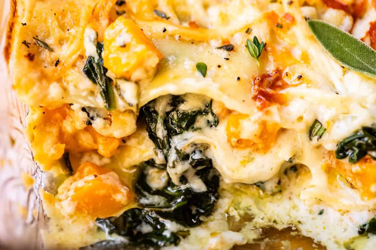 Close-up of a baked lasagna with layers of melted cheese, pasta, spinach, chunks of roasted butternut squash, and fresh herbs. The textures are creamy and golden, with visible leafy greens and orange squash.