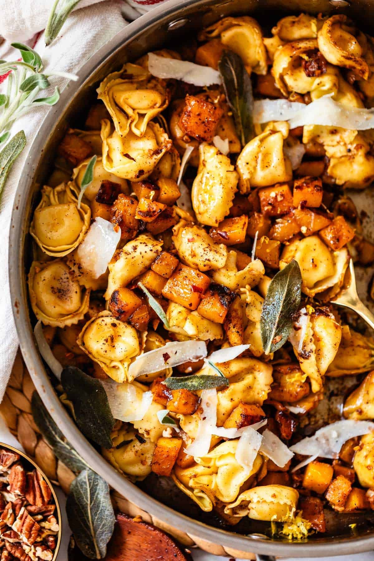 A close-up of a skillet filled with tortellini pasta, roasted butternut squash, crispy sage leaves, and shaved Parmesan cheese, all seasoned with black pepper.