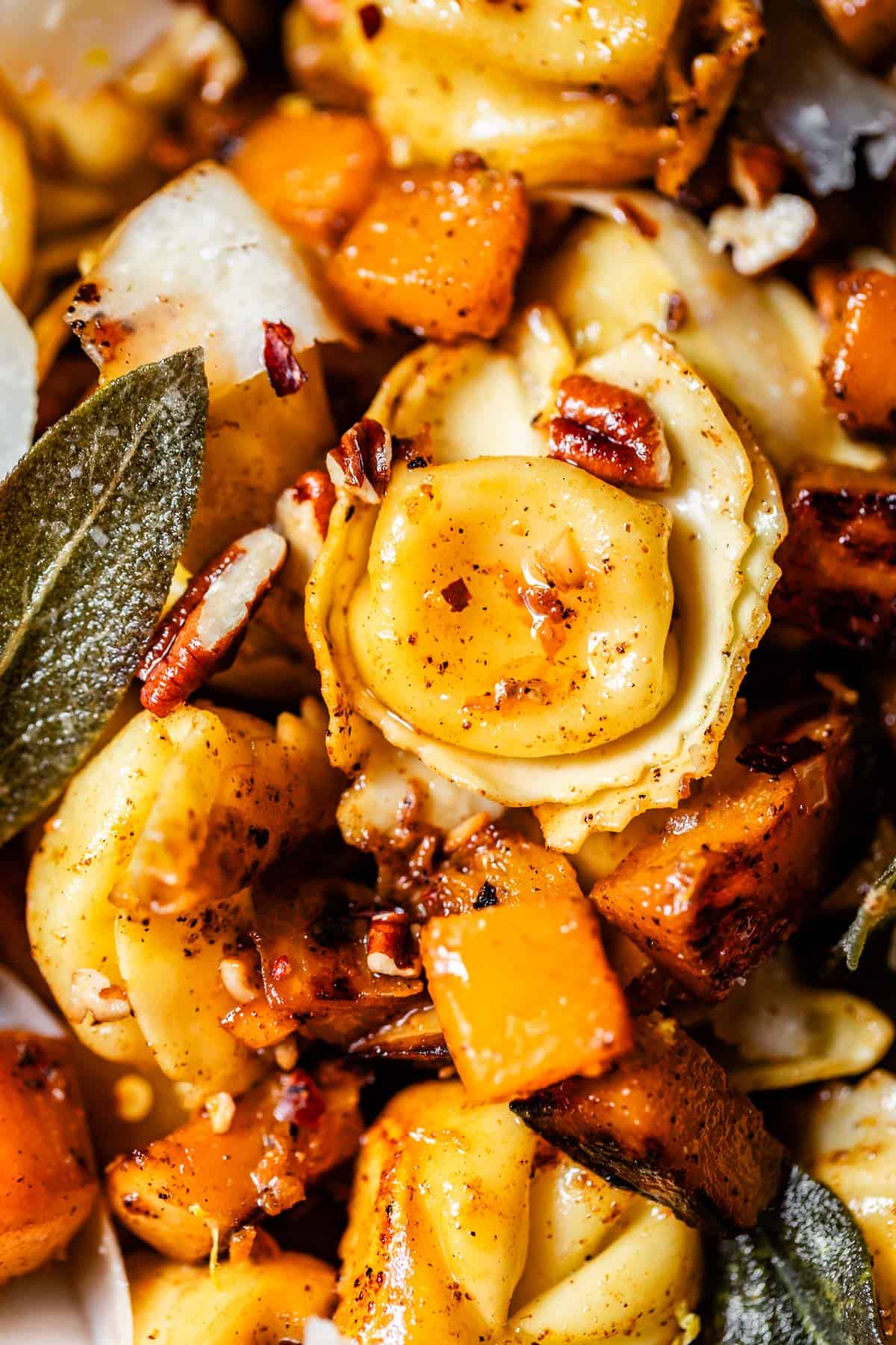 Close-up of tortellini pasta mixed with roasted butternut squash, pecans, and fresh sage leaves, all seasoned with spices. The dish looks warm, colorful, and garnished with crunchy nuts.