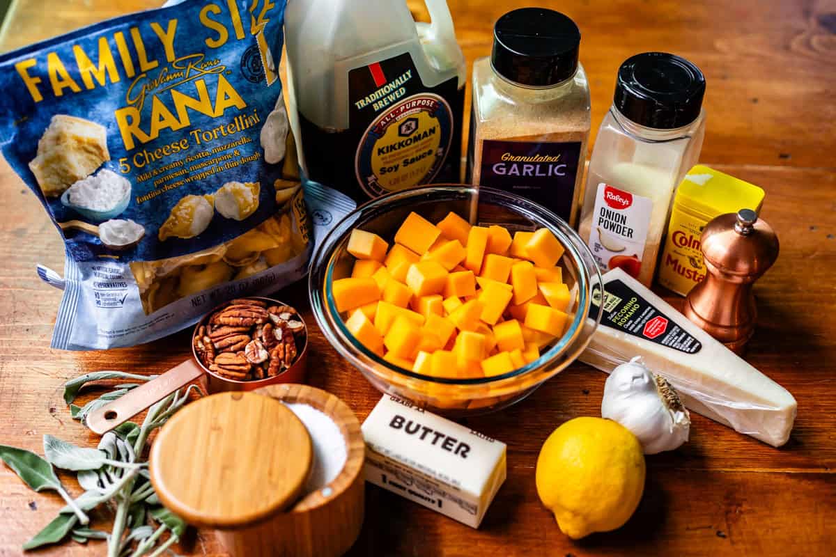 A variety of cooking ingredients on a wooden surface, including a bag of cheese tortellini, cubed butternut squash, butter, parmesan cheese, seasonings, soy sauce, lemon, pecans, and fresh sage leaves.