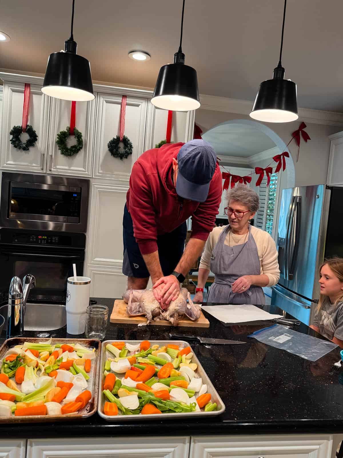 A man in a red hoodie stands on a kitchen counter, preparing a turkey while a woman and a girl watch. Trays of chopped vegetables are on the counter, and the kitchen is decorated for the holidays.