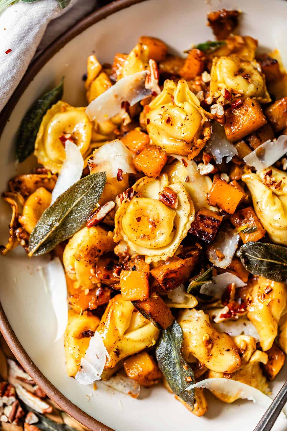 A close-up of a pasta dish featuring cheese-filled tortellini, roasted butternut squash, fresh sage leaves, shaved parmesan, and chopped nuts on a white plate. The meal looks vibrant and garnished with herbs.